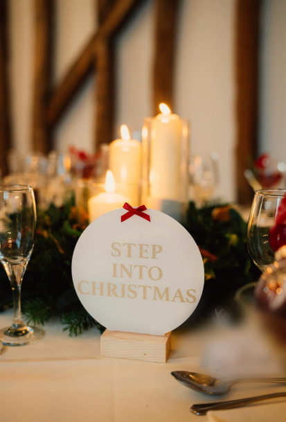 A round &ldquo;Step into Christmas&rdquo; table sign with a red bow among candles and pine