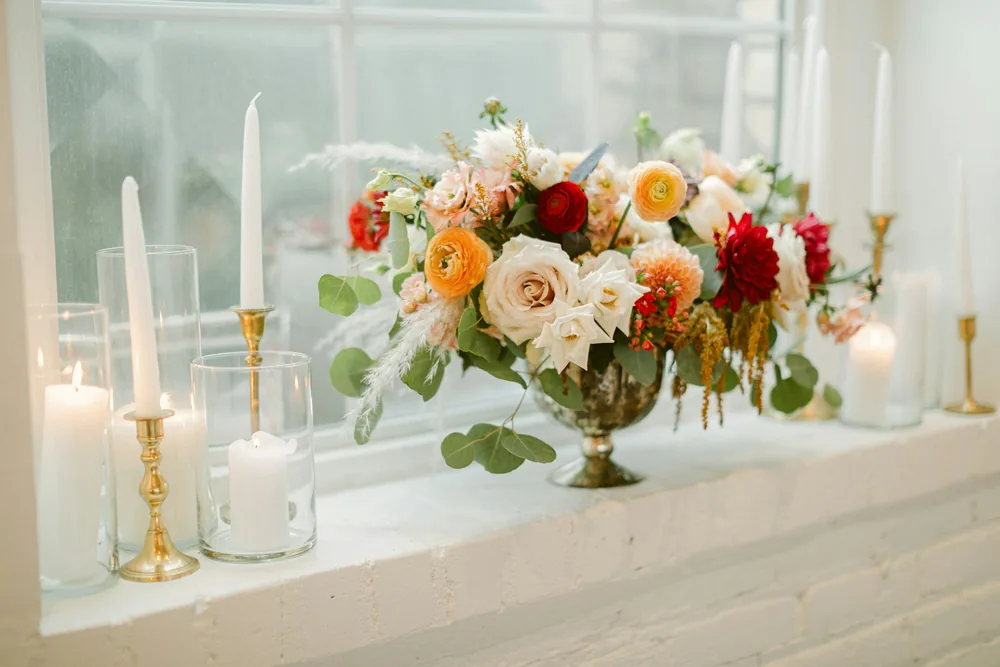 A floral arrangement of cream and red roses with tall candles on a windowsill