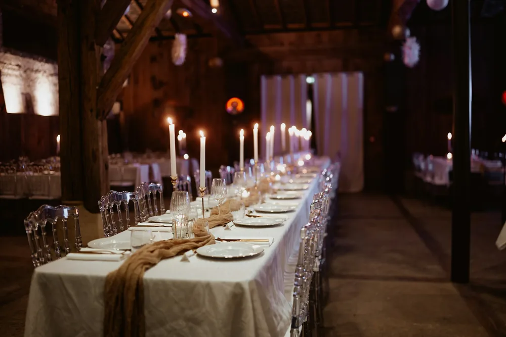 A long candlelit table dressed in white linen inside a dark timber barn